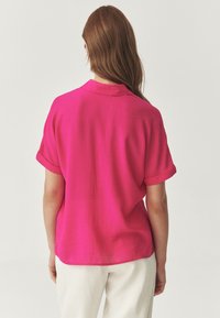 Camicia a maniche corte in rosa acceso, realizzata in tessuto strutturato, con colletto e polsini arrotolati. L'orlo è leggermente arrotondato sul retro.