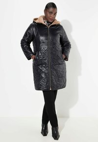 Zwarte gewatteerde pufferjas met een beige bontgevoerde capuchon, een ritssluiting vooraan en twee zijzakken, gestyled over zwarte aansluitende broek en laarzen.