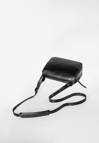 Schwarze Leder-Umhängetasche mit verstellbarem Riemen und Reißverschluss-Hauptfach, flach liegend auf einer weißen Oberfläche.