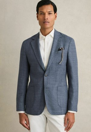 Grauer strukturierter Blazer mit zwei Fronttaschen, Schalkragen und einer einzigen Knopfschließe, kombiniert mit einem weißen Hemd und hellen Hosen.
