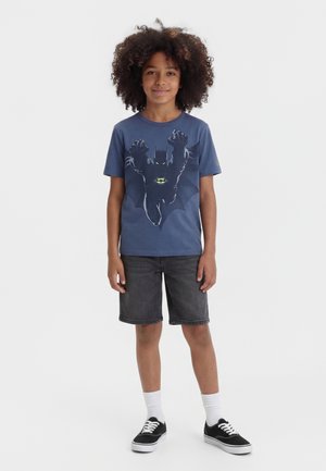 Enfant aux cheveux bouclés portant un t-shirt bleu avec un motif Batman, un short en jean noir, des chaussettes blanches et des baskets noires, debout sur un fond uni.