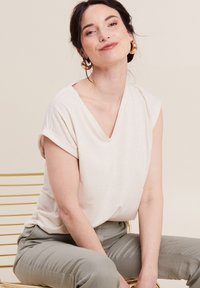 Helles beige Kurzarmoberteil mit V-Ausschnitt und subtiler Streifenstruktur. Getragen mit hellgrauen Hosen, sitzt auf einem goldenen Drahtstuhl.