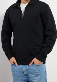 Hombre vestido con un pulóver negro de manga larga con cremallera frontal y bolsillo en el pecho, combinado con jeans de mezclilla azul claro, con las manos en los bolsillos.