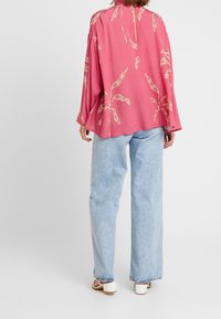 Långärmad rosa blus med beige bladmönster, löst passform, detaljerad öppen rygg. Matchas med ljusblå vida jeans och vita sandaler.