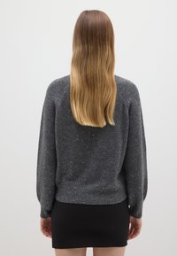 Pull en tricot gris à coupe ample, avec des accents de paillettes éparpillées et des poignets côtelés, porté sur une jupe noire.