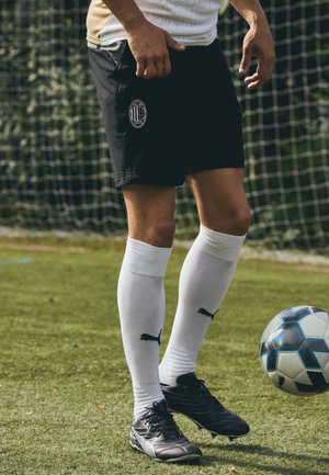 Schwarze Fußballshorts mit einem runden Logo, weiße knielange Socken mit schwarzem Logo und schwarze Fußballschuhe, die auf Gras neben einem Fußball stehen.