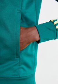 Turfärgad sweatshirt med känguruficka, ribbad mudd och gula randiga detaljer längs ärmen. Slät tyg med en mjuk textur.