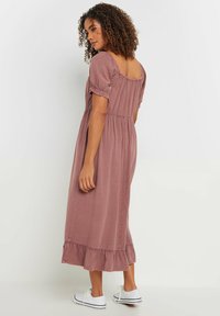 Vestido maxi de tela rosa polvo, con mangas abullonadas, dobladillo con volantes, y un corpiño ajustado con pliegues. Combinado con zapatillas blancas.