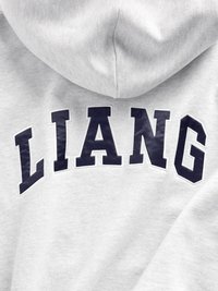 Hallitus hall vest, mille tagaküljel on suur tume sinine tikitud "LIANG". Puuvillane materjal on pehme tekstuuriga ning klassikalise kapuutsiga disain.