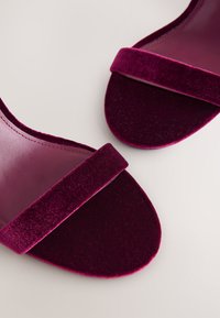 Pár bordó sametových sandálů s otevřenou špičkou a širokým páskem přes prsty, vystavených na světlém povrchu.