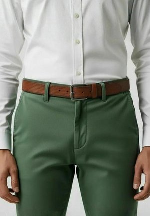Man wearing white button-up shirt tucked into green pants with a brown leather belt, standing with hands relaxed at sides.