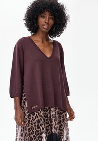 Bordowy sweter z dzianiny z rękawami do łokcia i dekoltem w serek, nałożony na przezroczystą spódnicę w panterkę z koronkowym akcentem.