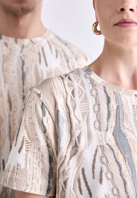 Beige mønstrede t-shirts med en struktureret design, der har grå og kremfarvede detaljer, rund halsudskæring og korte ærmer.