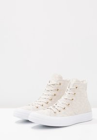 Beżowe sneakersy za kostkę z teksturowanej tkaniny, białą gumową podeszwą, złotymi oczkami i dopasowanymi sznurówkami. Posiadają wzorzysty design na cholewce.