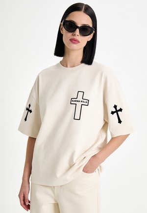 Vrouw met zwart bobkapsel en zonnebril, die een beige oversized T-shirt draagt met zwarte kruisgrafieken en "Karma Kills" tekst op de voorkant.