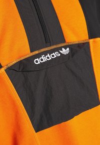 Pullover Adidas nero e arancione con cerniera a quarti, dotato di chiusura frontale con zip, pannelli a contrasto e logo bianco sulla tasca sul petto.