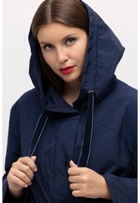 Laikaveden kestävä tummansininen takki, jossa on suuri huppu, etunapitus ja kiristysnarut. Sileä pinta ja kontrastiväriä kiristysnarujen yksityiskohdissa.