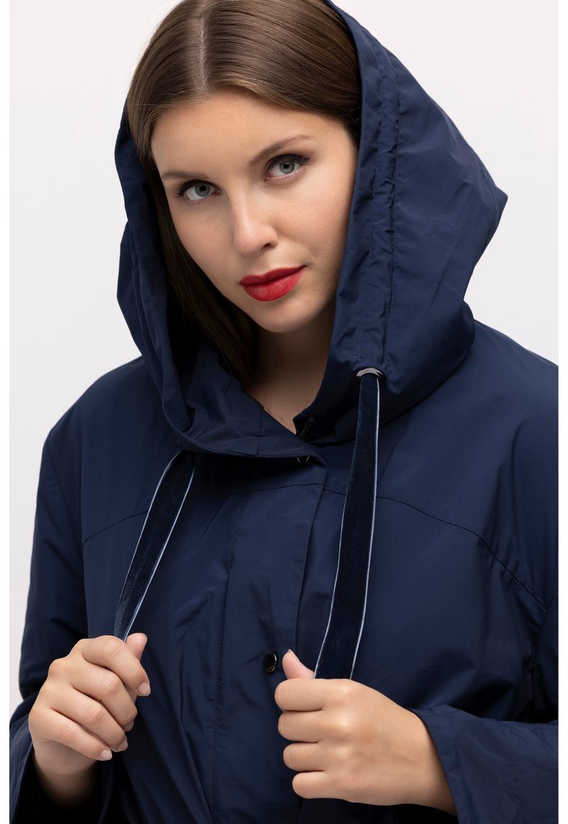 Laikaveden kestävä tummansininen takki, jossa on suuri huppu, etunapitus ja kiristysnarut. Sileä pinta ja kontrastiväriä kiristysnarujen yksityiskohdissa.