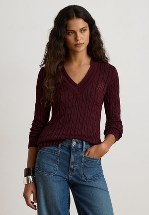 Femme aux longs cheveux bouclés foncés, portant un pull en maille torsadée bordeaux à col en V et un jean bleu, avec un bracelet en argent à son poignet gauche.