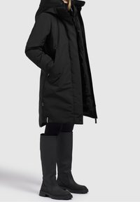 Parka nera impermeabile con cappuccio, dotata di cerniere e bottoni a pressione. Abbinata a stivali di gomma neri alti fino al ginocchio e leggings scuri.