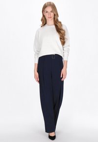 Jasnoszara dzianinowa sweter z długimi rękawami, w połączeniu z granatowymi spodniami o wysokim stanie z paskiem i plisowanym wzorem. Czarne buty.