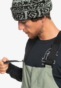 Bonnet tricoté vert foncé avec un motif noir, porté avec une chemise noire à manches longues et des salopettes vert clair avec des bretelles ajustables.
