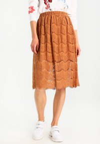 Lace midi nederdel i rustfarve med bølget kant, der har indviklede blomster mønstre og en elastisk talje. Kombineret med hvide sko.