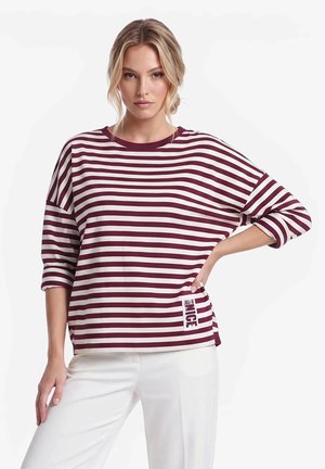 Donna con capelli biondi che indossa una maglia a maniche lunghe a righe bordeaux e bianche e pantaloni bianchi, in piedi con una mano sul fianco.