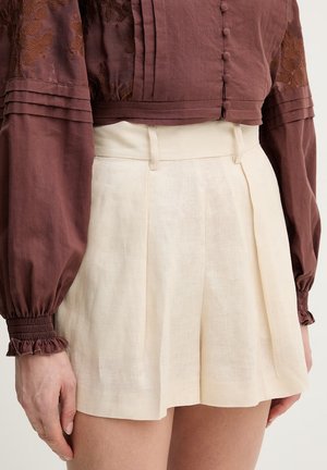 Pantaloncini in lino color crema a vita alta con pieghe e passanti per cintura indossati con una camicetta marrone a maniche lunghe con bottoni rivestiti in tessuto e polsini arricciati.