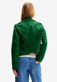 Veste courte en cuir synthétique vert avec un large col, des épaulettes et une texture lisse, associée à un jean bleu taille haute.