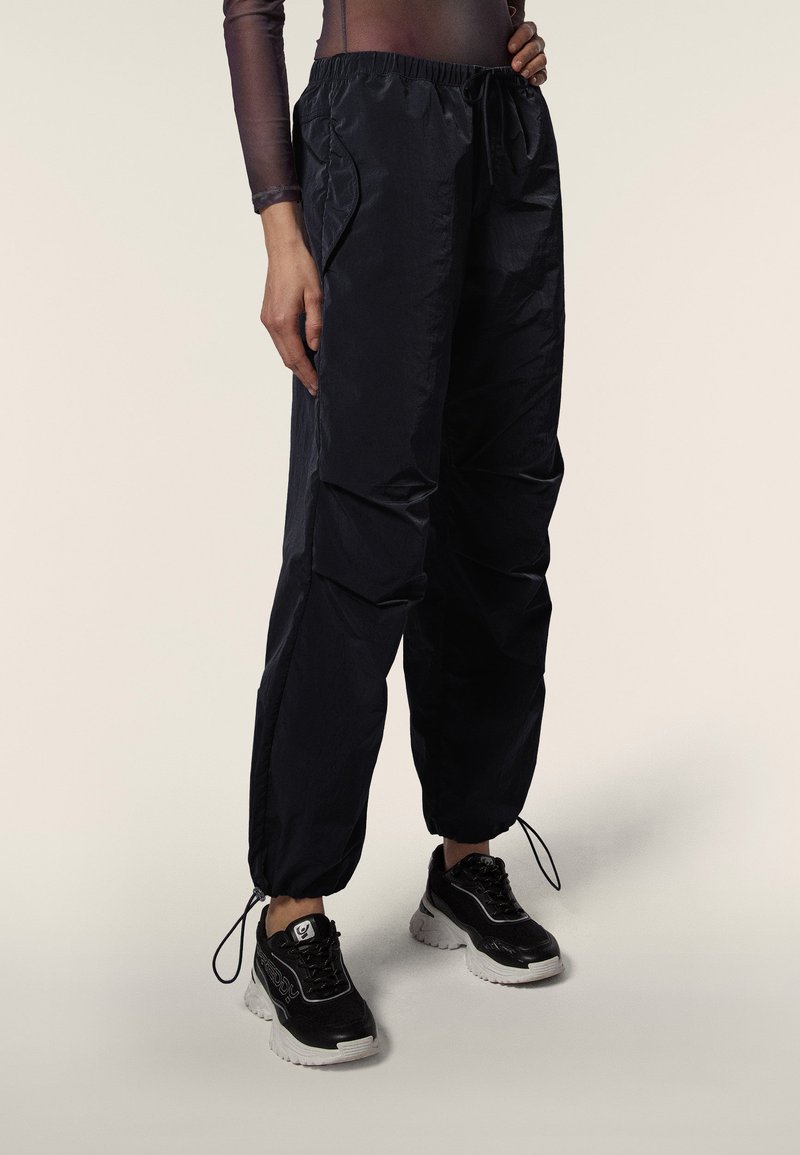 Person trägt schwarze Nylon-Hose mit Kordelzug und schwarze klobige Sneakers, steht vor einem schlichten hellen Hintergrund.