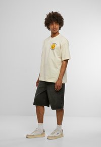 T-shirt en coton beige avec un graphique de smiley jaune circulaire, associé à un short en denim foncé et des baskets blanches avec des accents beiges.