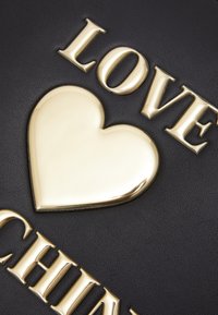 Musta tekstuurinen materiaali, jossa on kultaista kirjailua, joka lausuu "Rakkaus" ja kiiltävä kultainen sydämen aksentti. Suunnittelu on rohkea ja moderni.