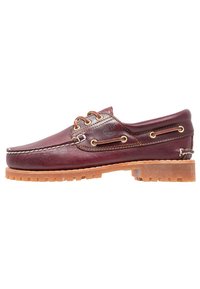 Chaussure bateau en cuir bordeaux avec semelle en caoutchouc, œillets dorés et finition texturée. Présente un logo sur le côté et des coutures détaillées.