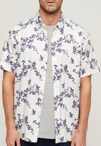 Camisa de botones de manga corta hecha de tejido ligero, con un fondo blanco y patrones florales morados, y un cuello estándar.