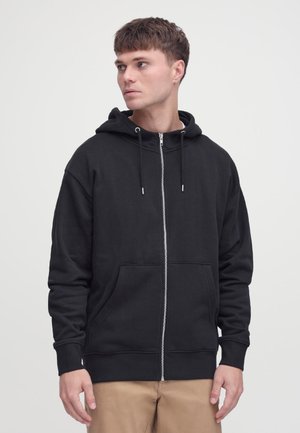 Giovane uomo che indossa una felpa con cappuccio nera con zip e tasche frontali, pantaloni color kaki, che guarda verso sinistra su uno sfondo semplice.