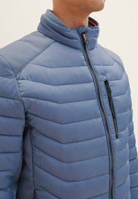 Pohodlná modrá bunda s vysokým golierom, ktorá má zipsovú náprsné vrecko a textúrované ramenné panely, vykazuje horizontálny prešívaný vzor.