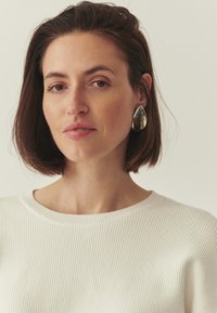 Pull blanc côtelé avec un col rond, présentant une texture lisse. De grandes boucles d'oreilles en forme de goutte argentées complètent le look.
