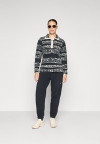 Genser med geometrisk svart og hvit mønster og høy krage, kombinert med svarte joggebukser. Modellen har på seg lyse joggesko og solbriller.