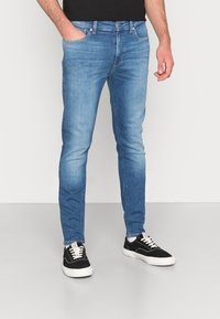 Světle modré džínové kalhoty s úzkým střihem, pětikapsovým designem a jemným vyblednutím. Doplněné černými teniskami s bílými tkaničkami.