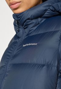 Mørkeblå dynejakke med høj krave, quiltet design og hvidt "Peak Performance"-logo på den øverste brystdel. Teksturen virker glat.