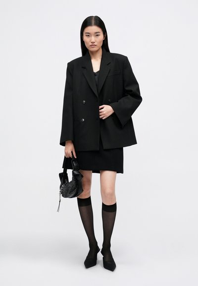 Črn oversized blazer z dvojnimi gumbi, v kombinaciji s črno mini krilom in črnimi nogavicami do kolen. Drži teksturirano črno torbico.