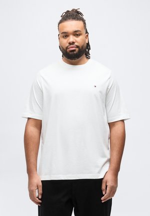 Mand med dreadlocks og skæg iført en almindelig hvid kortærmet T-shirt og sorte bukser, der står mod en lys baggrund.