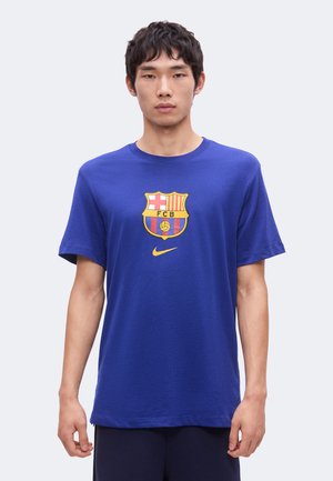 Jeune homme portant un t-shirt bleu avec le logo du FC Barcelone et le swoosh jaune de Nike à l'avant, debout devant un fond clair uni.