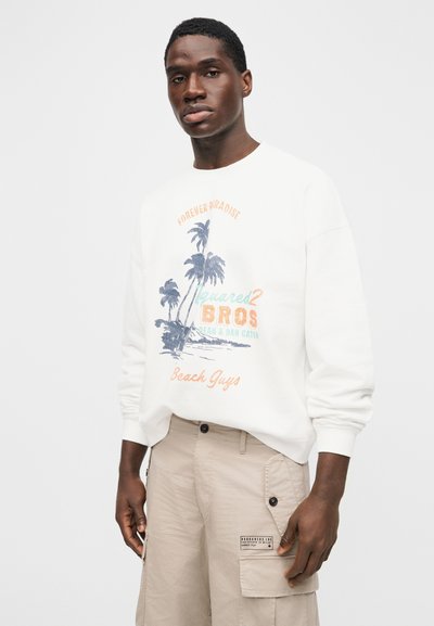 Νέος άνδρας που φοράει λευκή μπλούζα με σχέδιο από φοίνικες και μπεζ παντελόνι cargo, στέκεται μπροστά σε απλό ανοιχτό φόντο.