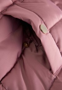 Acercamiento de un bolsillo acolchado de una chaqueta rosa con un botón de presión metálico brillante parcialmente abierto que muestra el forro interior de tela.