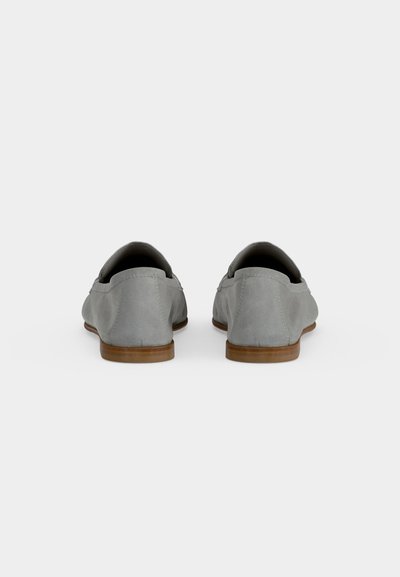 Paire de chaussures slip-on en daim gris avec semelles plates marron, vues de dos sur un fond clair uni.