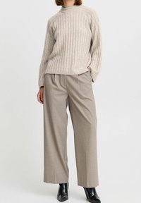 Jersey beige con relieve y cuello alto, combinado con pantalones de pierna ancha a rayas finas en marrón claro. La modelo lleva botas negras hasta el tobillo.