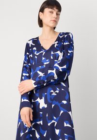 Μακριά μανίκια φόρεμα σε σκούρο μπλε χρώμα με αφηρημένα μπλε και λευκά σχέδια, διαθέτοντας V-λαιμόκοψη και λεπτομέρειες με κουμπιά στα μανίκια. Λείο ύφασμα.
