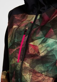 Veste à motifs géométriques multicolores avec manches noires, une fermeture éclair à l'avant et une poche verticale zippée rose vif sur la poitrine.